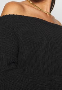 Femme portant un pull noir en maille à épaules dénudées et un collier chaîne doré, montrant ses épaules et son haut de poitrine nus.