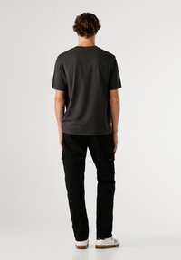 Tricou scurt cu mâneci negre, din bumbac, cu un croi lejer. Purtați cu pantaloni cargo negri și adidași albi, stând pe un fundal simplu.