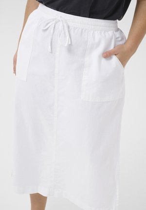 Naine kannab valget põlveni ulatuvat seelikut, millel on ees taskud ja nööriga vöökoht, parem käsi paremas taskus, musta topiga.