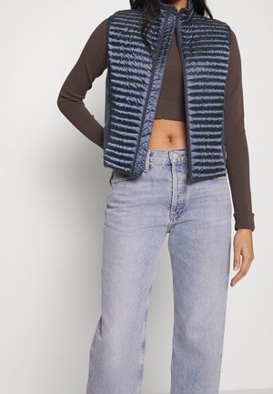 Persoon draagt een donker cropped donsjack over een bruine longsleeve en lichtblauwe high-waisted jeans, staand tegen een effen achtergrond.