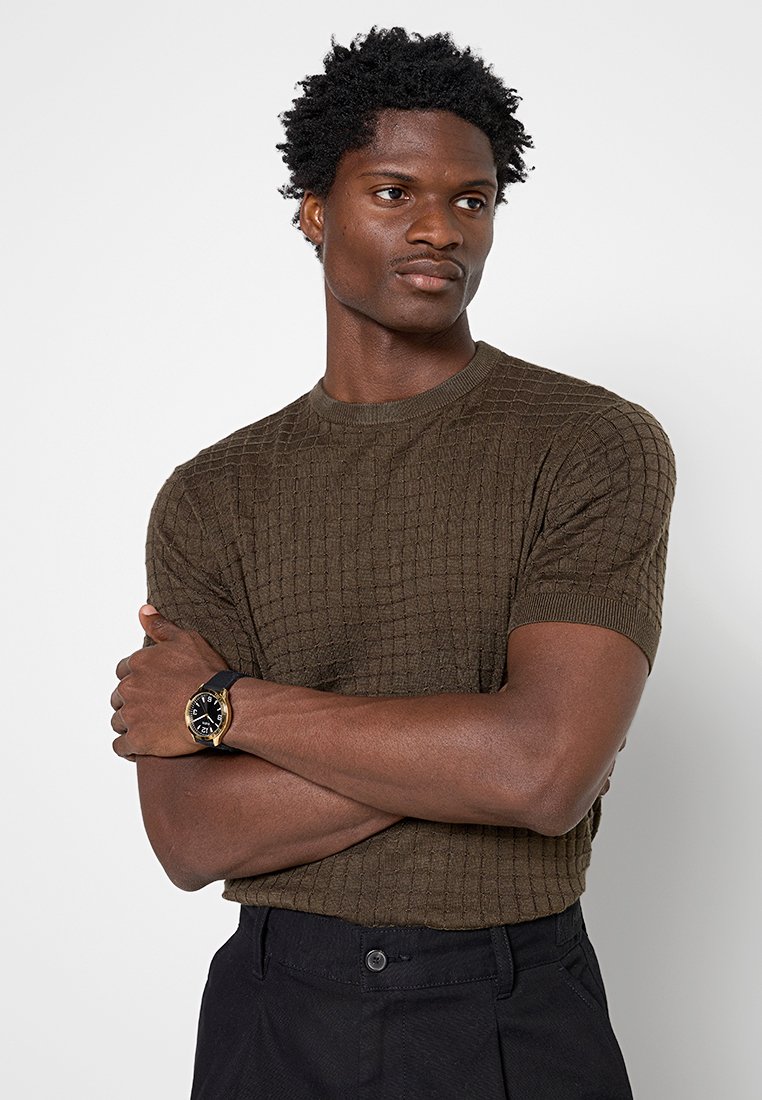 Hombre con los brazos cruzados que lleva un suéter de manga corta texturizado de color verde oliva, pantalones negros y un reloj negro con bisel dorado, mirando hacia la derecha.