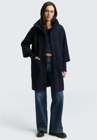 Marineblaues übergroßes Coat aus strukturiertem Stoff, mit weitem Kragen und zwei Fronttaschen, kombiniert mit weiten Jeans und schwarzen Stiefeln.