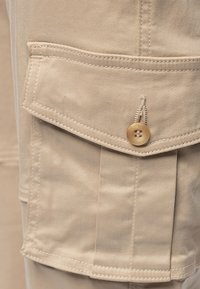Beige cargo byxor i tyg med en sidoficka, detaljerad sömnad och en fyrhålsknapp. Materialet är mjukt med en lätt texturerad yta.