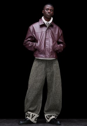 Tânăr cu mâinile în buzunare, purtând o geacă de piele bordo, pantaloni largi din lână gri cu tivuri întoarse care afișează text negru și pantofi negri.