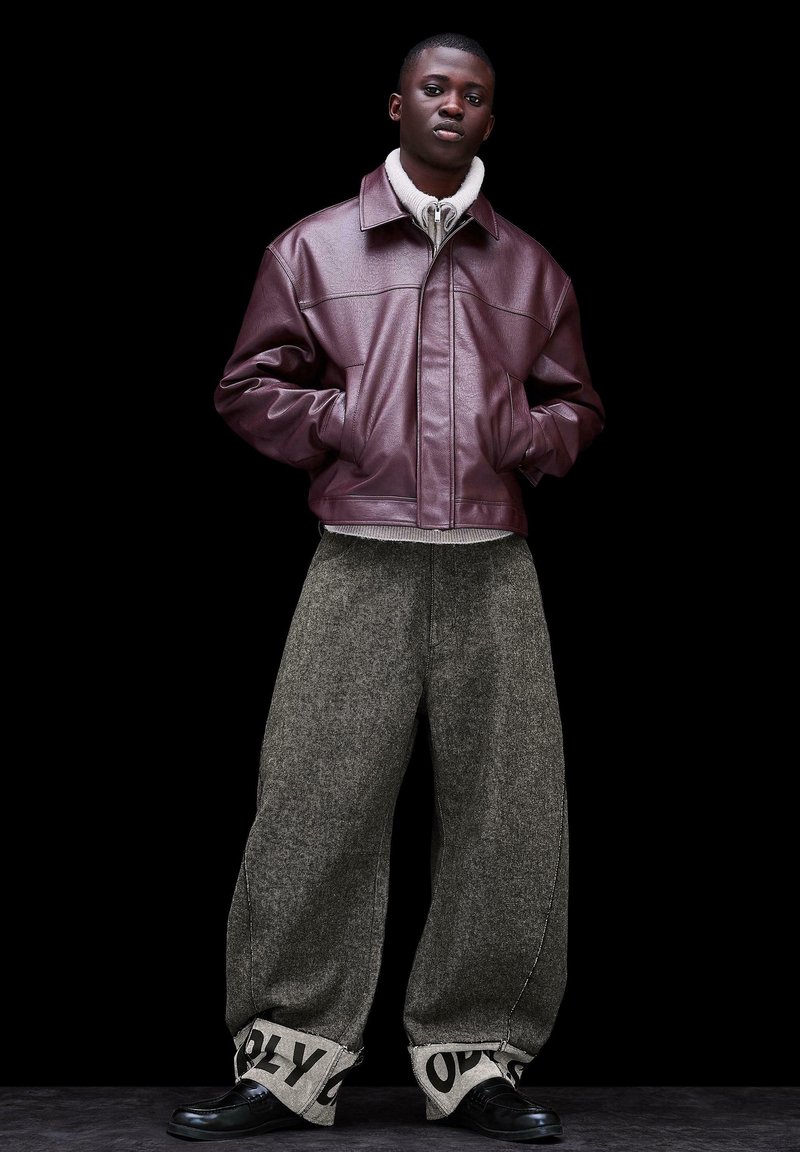 Junger Mann mit Händen in den Taschen, der eine kastanienbraune Lederjacke, graue, weit geschnittene Wollhosen mit aufgekrempelten Säumen und schwarzer Schrift sowie schwarze Schuhe trägt.