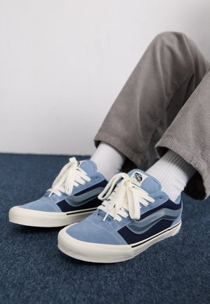 Blaue Wildledersneaker mit navy Akzenten, weißen Schnürsenkeln und einem auffälligen Streifenmuster. Strukturiert Oberfläche und Gummisohle, getragen mit weißen Socken und grauen Hosen.