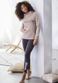 Beige Kapuzenpullover mit Fronttasche und "Bench"-Logo; kombiniert mit dunklen Leggings, die das "Bench"-Logo an der Seite tragen. Barfuß. Sanfte Texturen.