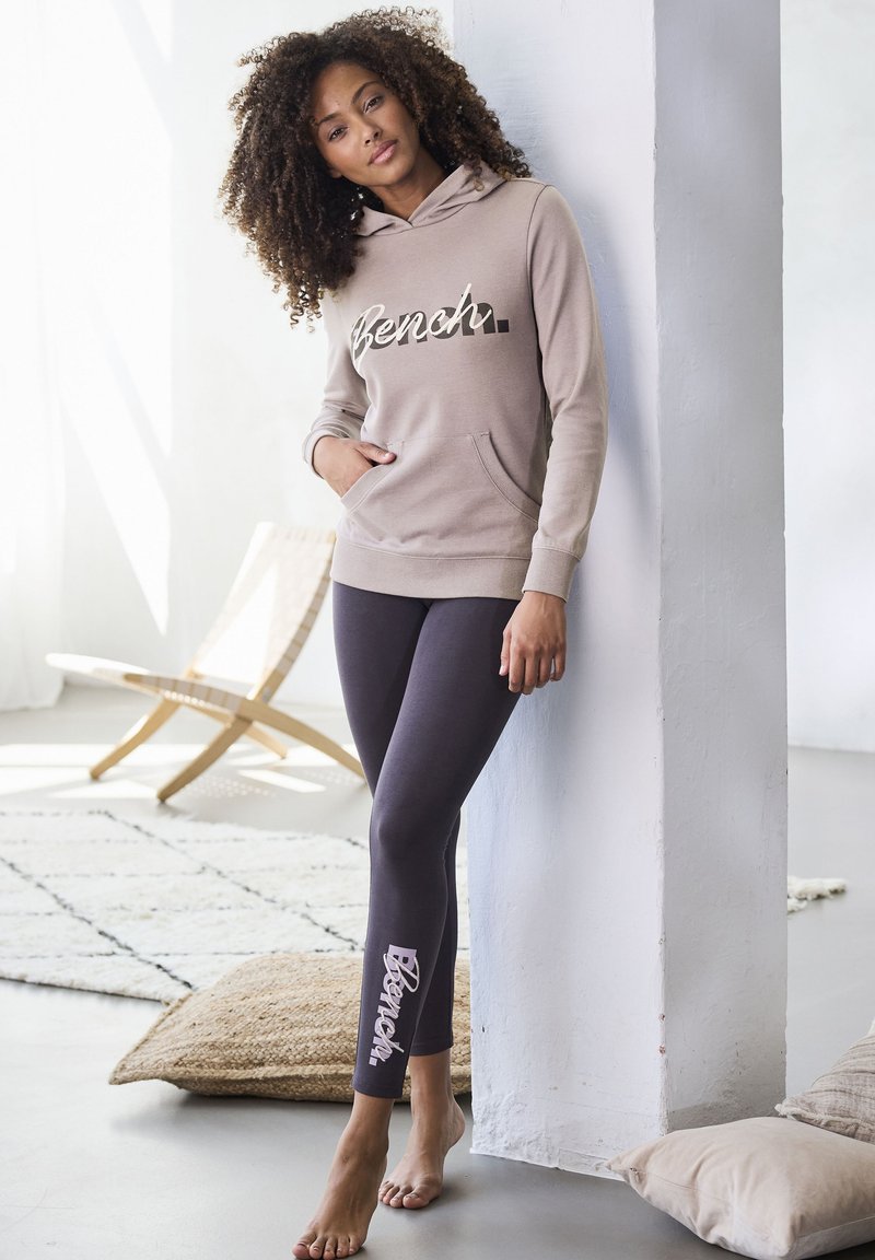 Beige Kapuzenpullover mit Fronttasche und "Bench"-Logo; kombiniert mit dunklen Leggings, die das "Bench"-Logo an der Seite tragen. Barfuß. Sanfte Texturen.