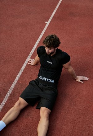 Un hombre vestido con ropa deportiva negra está sentado en una pista de atletismo roja, apoyándose hacia atrás sobre las manos junto a una línea blanca que delimita una calle.