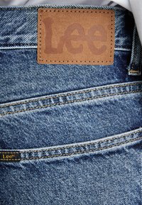 Denim jeans with a brown leather label featuring the embossed text "Lee." Blue wash with visible stitching details and pocket outline.