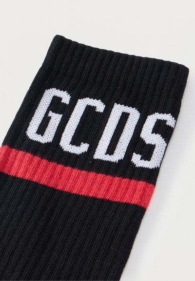 Μαύρη ριγωτή κάλτσα με λευκά γράμματα GCDS και μία οριζόντια κόκκινη γραμμή κάτω από το κείμενο.