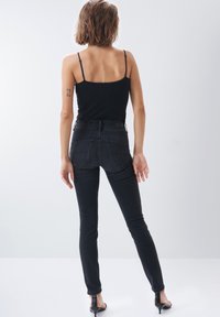 Jeans ajustados negros de tejido vaquero con una cintura alta. Los bolsillos traseros presentan costuras con una textura sutil. El modelo lleva una camiseta negra sin mangas y tacones.