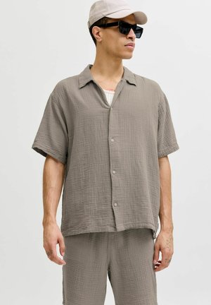 Mann trägt hellgraues kurzärmeliges Hemd mit Knöpfen und passende Shorts, beige Mütze und schwarze rechteckige Sonnenbrille, steht vor weißem Hintergrund.