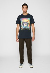 Jūras flotes kokvilnas t-krekls ar krāsainu grafiku, kurā attēlots šimpanzes tēls ar austiņām un uzraksts "Cyber Chimp". Apvienots ar tumši brūnām biksēm.