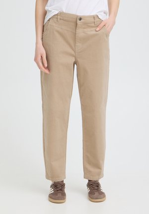 Persoon draagt beige rechte broek met zijzakken en bruine vetersneakers, staand tegen een effen achtergrond.