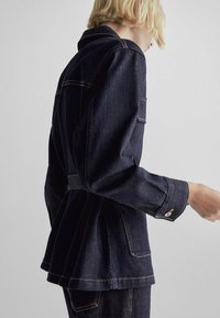 Ciemna kurtka dżinsowa o dopasowanym kroju, z dużymi kieszeniami i mankietami zapinanymi na guziki. Posiada kontrastowe przeszycia oraz pasek w talii dla podkreślenia sylwetki.