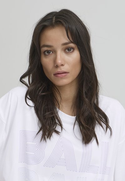 Bijela majica s ljubičastim obrubom teksta "BALL." Ima opušteni kroj i okrugli izrez, nosi je model s dugom smeškom.