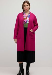 Langer, magentafarbener Cardigan mit offenem Frontbereich, zwei Taschen, getragen über einem mehrfarbigen gemusterten Hemd, kombiniert mit einem schwarzen gefalteten Rock und Stiefeletten.