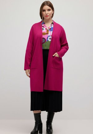 Long cardigan magenta avec ouverture sur le devant, deux poches, porté par-dessus une chemise à motifs multicolores, associé à une jupe plissée noire et des bottines.