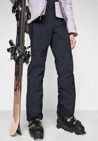 Smučarske hlače iz temno modrega blaga, ki imajo sproščen kroj, ojačana kolena in vodoodporno teksturo, v kombinaciji z black smučarskimi čevlji in desko za snowboard.