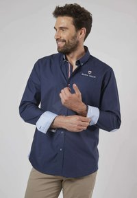 Chemise bleu marine à manches longues avec des poignets et un col en bleu clair contrastant. Présente un logo brodé et une poche poitrine. Tissu lisse.