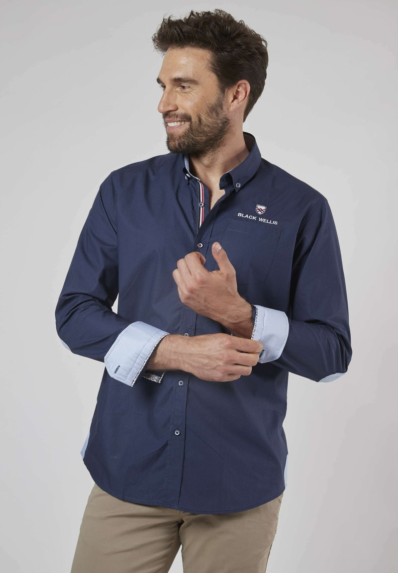Chemise bleu marine à manches longues avec des poignets et un col en bleu clair contrastant. Présente un logo brodé et une poche poitrine. Tissu lisse.