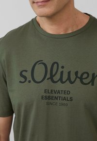 Oliwkowa bawełniana koszulka z czarnym napisem: "s.Oliver ELEVATED ESSENTIALS SINCE 1969" z przodu, z okrągłym dekoltem.