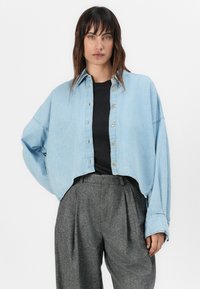 Chemise en denim bleu clair coupée avec un devant à boutons, coupe ample et manches longues, portée sur une chemise noire et un pantalon gris plissé.