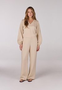 Beige twee-delig kledingstuk met een v-hals, lange mouwen met elastische manchetten, en wijdvallende broek met een relaxed fit, gemaakt van lichte stof.