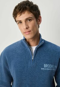 Mėlynas polarinis megztinis su pusiau užtrauktuku, tekstūruotas audinys ir šviesiai mėlynu užrašu "BROOKLYN WILLIAMSPORT" priekyje.
