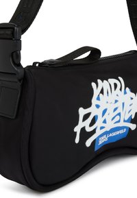 Černá nylonová taška přes rameno s kulatým tvarem, zdobená bílým textem ve stylu graffiti a modrou akcentovou nálepkou. Obsahuje nastavitelné ramenní popruhy a zip.