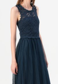 Námořnická modrá šaty s krajkovým korzetem a květinovými detaily, s průhlednou tylovou sukní. Bez rukávů s kulatým výstřihem.