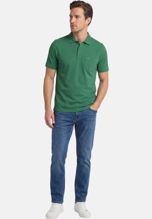 Man draagt een groen poloshirt met korte mouwen en een zakje, blauwe spijkerbroek en witte sneakers, staat met één hand in de zak en kijkt opzij.