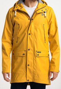 Man som bär en gul regnjacka med huva, framzipper och tryckknappar, fyra framfickor och vita dragsnören på huvan.