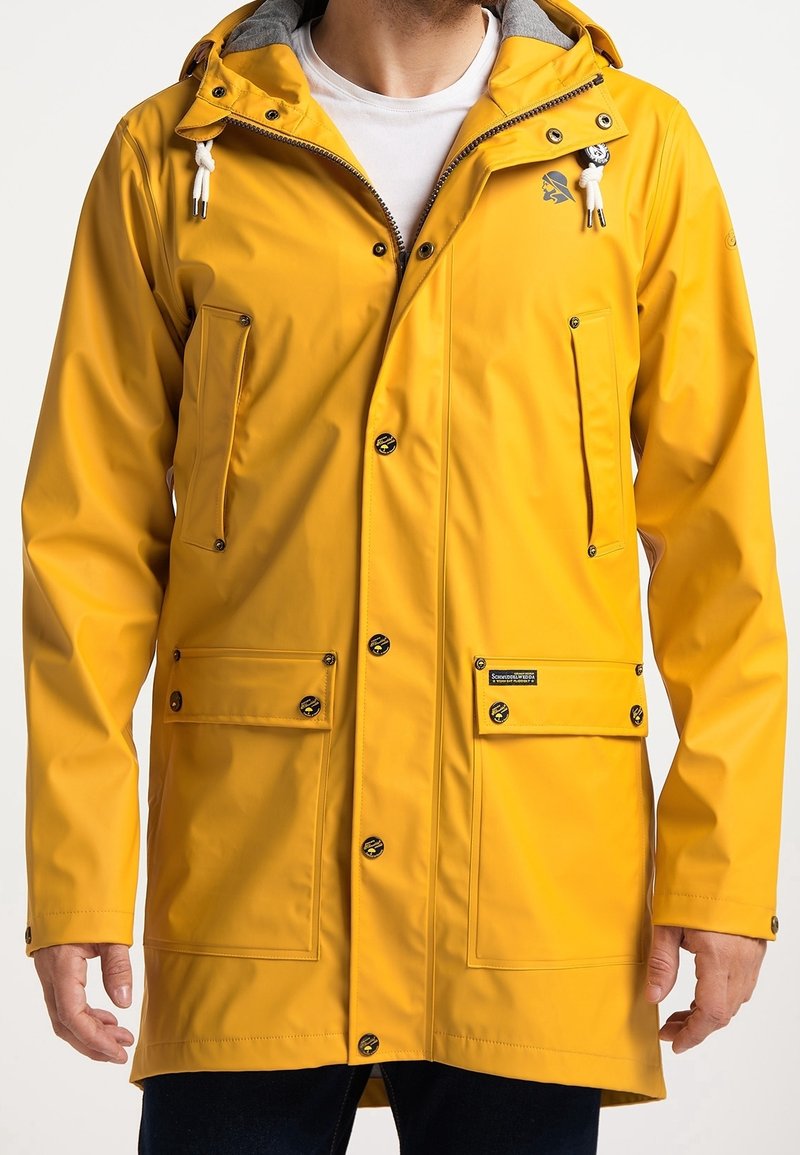 Man som bär en gul regnjacka med huva, framzipper och tryckknappar, fyra framfickor och vita dragsnören på huvan.
