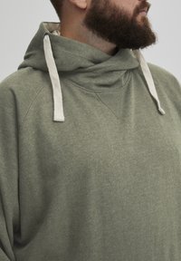 Sudadera gris-verde de corte relajado, con una gran capucha caída y cordones de color crema. Tejido suave con una ligera textura.
