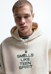 Beige hoodie met de tekst "SMELLS LIKE TEEN SPIRIT" in donkergroen. Voorzien van een zilveren ketting en een gestreept shirt eronder.