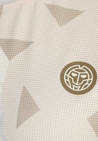 Tela texturizada en beige claro con patrones triangulares en tonos sutiles. Presenta un logo redondo en marrón con un diseño de cara estilizada.