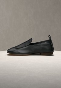Μαύρο δερμάτινο παπούτσι τύπου slip-on με επίπεδη σόλα, στρογγυλεμένη μύτη και χαμηλό τακούνι πάνω σε ουδέτερη επιφάνεια με γκρι φόντο.