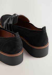 Juoda suede avalynė su blizgiu juodu dirželiu, apvaliu toe ir storu platformos padu su rudu akcentu apačioje.