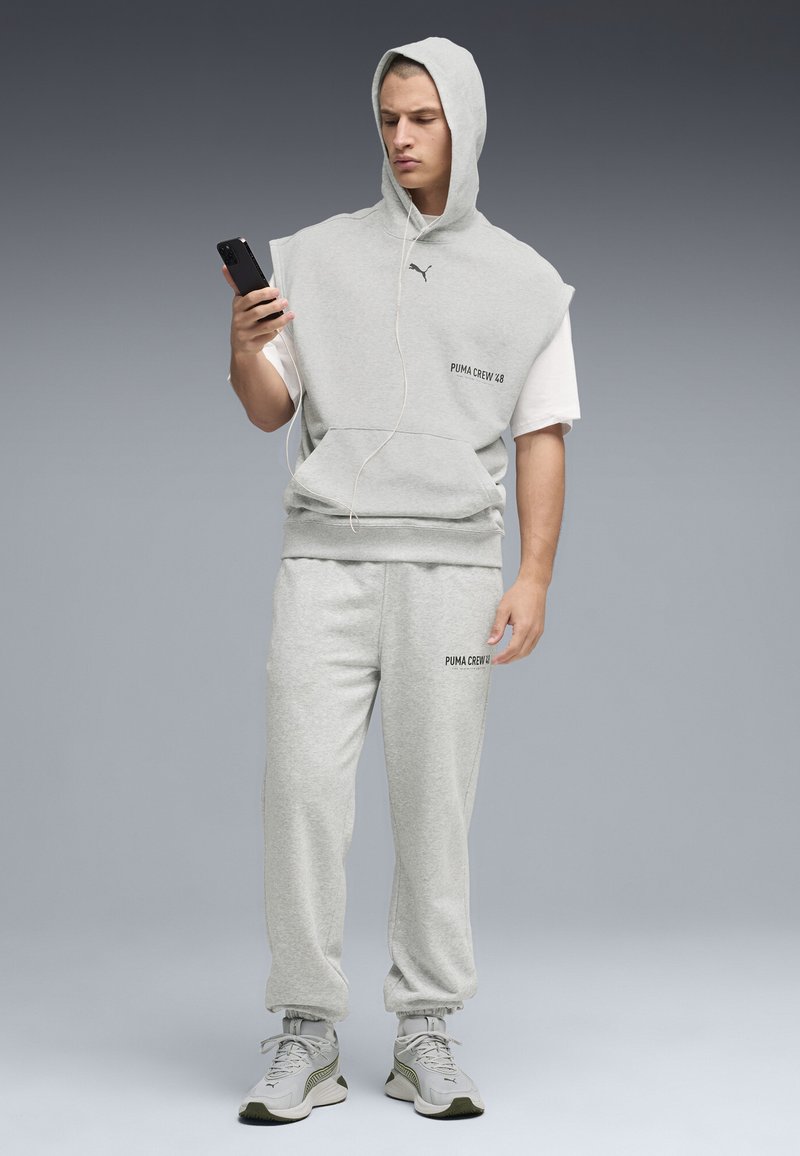 Άνδρας με γκρι αμάνικο φούτερ με κουκούλα και φόρμα, φοράει λευκά αθλητικά παπούτσια και ακουστικά, κοιτάζοντας το κινητό του μπροστά σε γκρι φόντο.