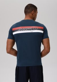 Camiseta azul marino con cuello redondo. Rayas horizontales en rojo, blanco y negro en la parte superior de la espalda. "CRAFTED GOODS" impreso en gris.