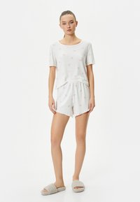 Vitt bomullsloungewear-set med en kortärmad topp prydd med små rosa blommönster, i kombination med matchande shorts och grå mysiga tofflor.