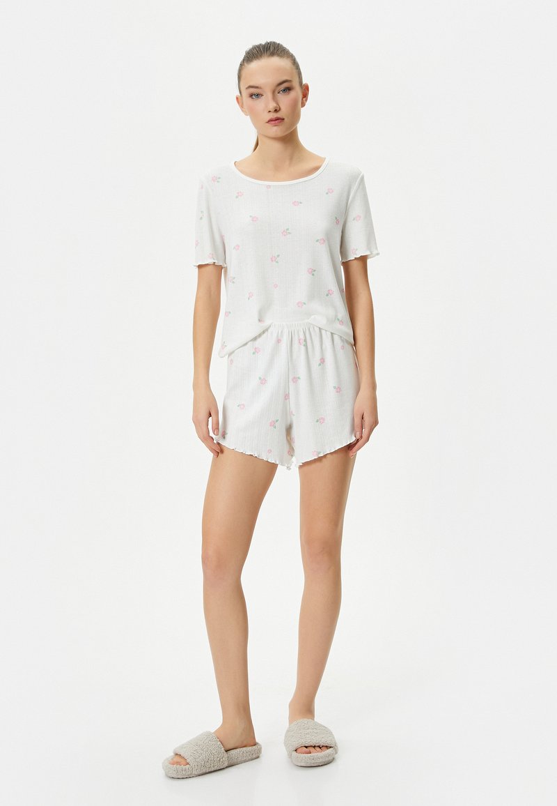 Vitt bomullsloungewear-set med en kortärmad topp prydd med små rosa blommönster, i kombination med matchande shorts och grå mysiga tofflor.