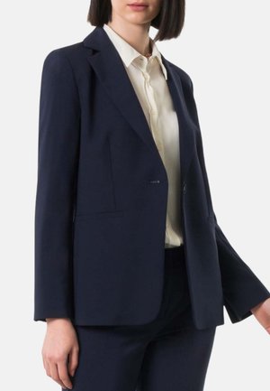Donna che indossa un blazer blu navy e pantaloni abbinati sopra una camicia crema con bottoni, in piedi davanti a uno sfondo chiaro e neutro.