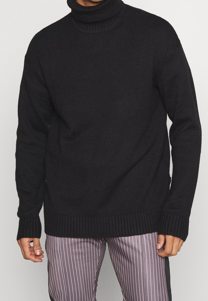 Schwarzer gestrickter Rollkragenpullover mit geripptem Kragen und Bündchen, lockerer Passform und weicher Textur. Kombiniert mit gestreiften Hosen.