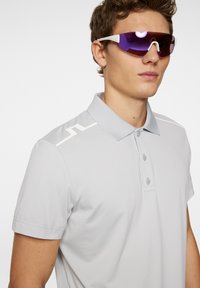 Light gray polo shirt with a three-button placket, featuring white accents on the shoulders. Worn with white-framed purple sunglasses.