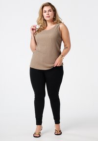 Tan geribbelde tanktop met een ronde hals, gedragen met zwarte strakke broek. Model heeft gouden sieraden en zwarte hoge hakken aan.