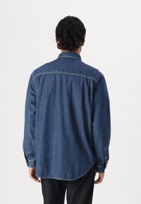 Camisa de mezclilla azul con mangas largas, puños abotonados, cuello extendido y dobladillo curvado. Detalles de costura visibles a lo largo de las costuras.