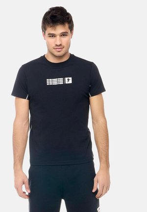 Giovane uomo con T-shirt nera con testo bianco che recita "WE ARE COMBAT SPORTS" e pantaloni neri coordinati, in piedi davanti a uno sfondo semplice.
